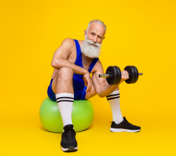 Die Abbildung zeigt einen durchtrainierten älteren Mann, der vor einem gelben Hintergrund auf einem grünen Gymnastikball sitzt und eine Hantel in der Hand hält